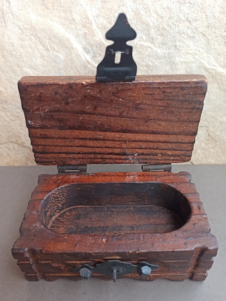 May include: A small wooden box with a hinged lid and a metal latch. The box is made of dark wood and has a simple design. The inside of the box is empty.