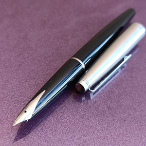 Könnte beinhalten: Ein schwarzer Füllfederhalter mit silberner Kappe und Feder, auf einer violetten Oberfläche. Die Kappe ist abgenommen und zeigt die Feder. Der Stift hat einen silbernen Ring.