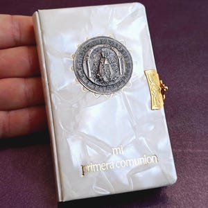 May include: A small, pearlescent white book with a silver medallion and gold clasp. The medallion features a religious figure. The words "mi primera comunion" are printed in gold on the cover. The book is held in a hand.