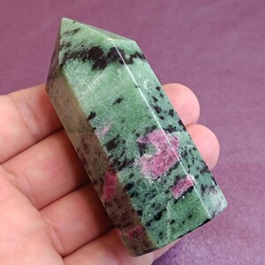 May include: A polished gemstone obelisk. The stone is a mix of green, black, and pink hues. The obelisk is held in a hand, showcasing its faceted sides and pointed top. The gemstone's colors and patterns are clearly visible.