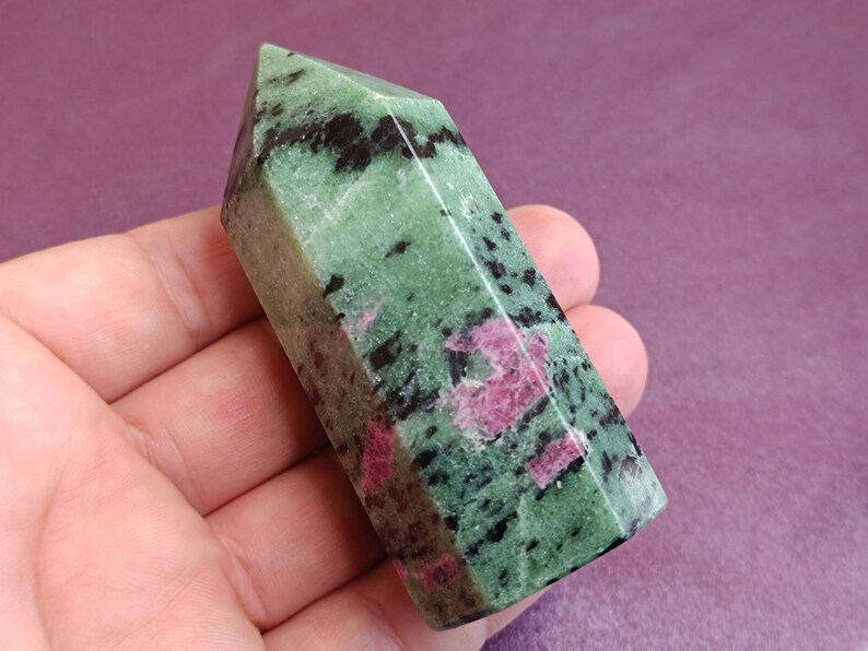 May include: A polished gemstone obelisk. The stone is a mix of green, black, and pink hues. The obelisk is held in a hand, showcasing its faceted sides and pointed top. The gemstone's colors and patterns are clearly visible.