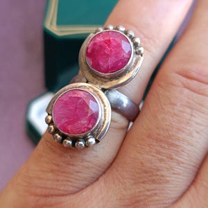 Puede incluir: Un anillo de plata con dos grandes piedras preciosas redondas, facetadas y de color rosa intenso. El anillo tiene un dise&ntilde;o &uacute;nico con un engaste de bisel y peque&ntilde;os detalles de cuentas alrededor de las piedras. El anillo est&aacute; en un dedo.