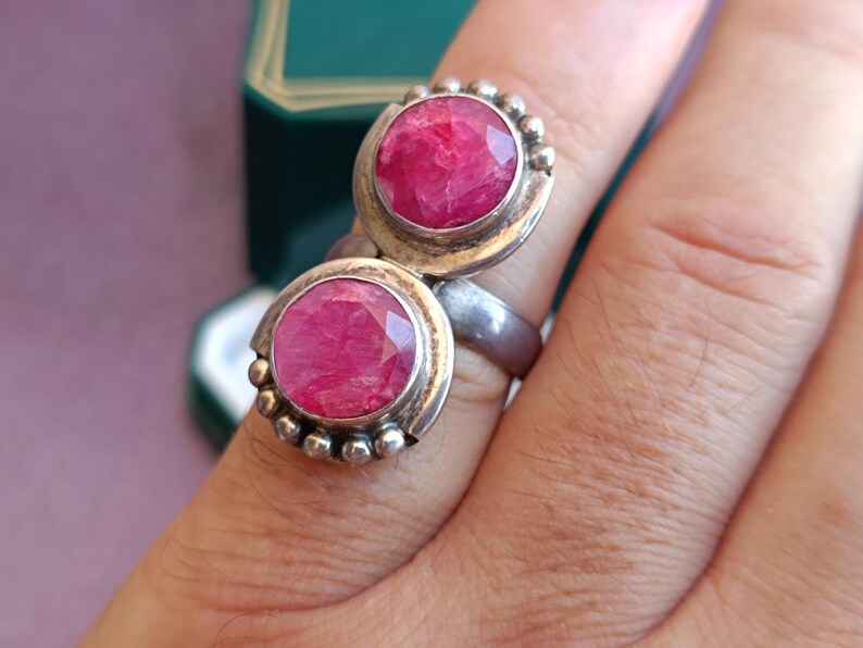 Puede incluir: Un anillo de plata con dos grandes piedras preciosas redondas, facetadas y de color rosa intenso. El anillo tiene un dise&ntilde;o &uacute;nico con un engaste de bisel y peque&ntilde;os detalles de cuentas alrededor de las piedras. El anillo est&aacute; en un dedo.