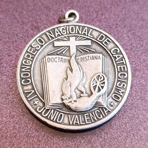 Peut inclure: Un pendentif rond argenté avec l'inscription "VI CONGRESO NACIONAL DE CATECISMO" et "JUNIO VALENCIA" sur le bord. Le centre représente une croix, une flamme, un livre ouvert avec l'inscription "DOCTRINA CRISTIANA" et un symbole religieux.