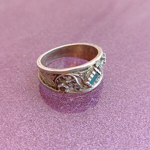 May include: A silver-toned metal ring featuring an intricate openwork design with floral-inspired patterns and a small, rectangular, dark green inlay set into the band. The metal surface shows a weathered, textured finish throughout the band.