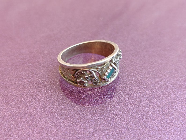 May include: A silver-toned metal ring featuring an intricate openwork design with floral-inspired patterns and a small, rectangular, dark green inlay set into the band. The metal surface shows a weathered, textured finish throughout the band.