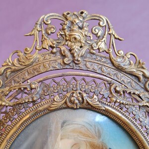 K&ouml;nnte beinhalten: Antiker, goldfarbener, ovaler Bilderrahmen mit aufwendigen floralen und scrollartigen Designs. Der Rahmen umgibt ein Portr&auml;t einer Frau mit blondem Haar. Der Rahmen befindet sich vor einem lila Hintergrund.