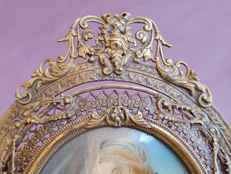 K&ouml;nnte beinhalten: Antiker, goldfarbener, ovaler Bilderrahmen mit aufwendigen floralen und scrollartigen Designs. Der Rahmen umgibt ein Portr&auml;t einer Frau mit blondem Haar. Der Rahmen befindet sich vor einem lila Hintergrund.