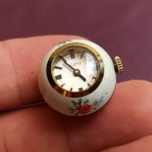 May include: A small, vintage watch with a white, spherical case. The watch face is silver with black hands and hour markers. The case is adorned with floral accents in pink, blue, and green. The watch has a gold-colored bezel and crown. The word "Ilana" is printed on the watch face.