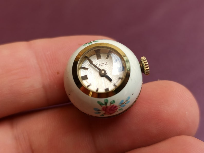May include: A small, vintage watch with a white, spherical case. The watch face is silver with black hands and hour markers. The case is adorned with floral accents in pink, blue, and green. The watch has a gold-colored bezel and crown. The word "Ilana" is printed on the watch face.