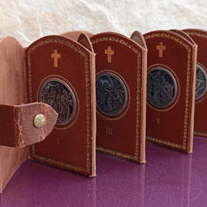 Pode incluir: Um conjunto de livros religiosos em couro castanho com medalhões de metal em relevo e detalhes dourados. Cada livro apresenta uma cruz na capa e é preso com uma tira de couro e fecho de botão. Os livros estão expostos numa superfície roxa.