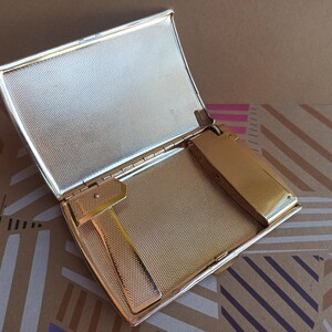 May include: A gold-toned cigarette case with a built-in lighter. The case is open and the lighter is visible. The case has a textured surface.