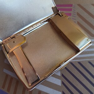 May include: A gold-colored metal cigarette case with a built-in lighter. The case is open and the lighter is visible. The case has a textured surface.