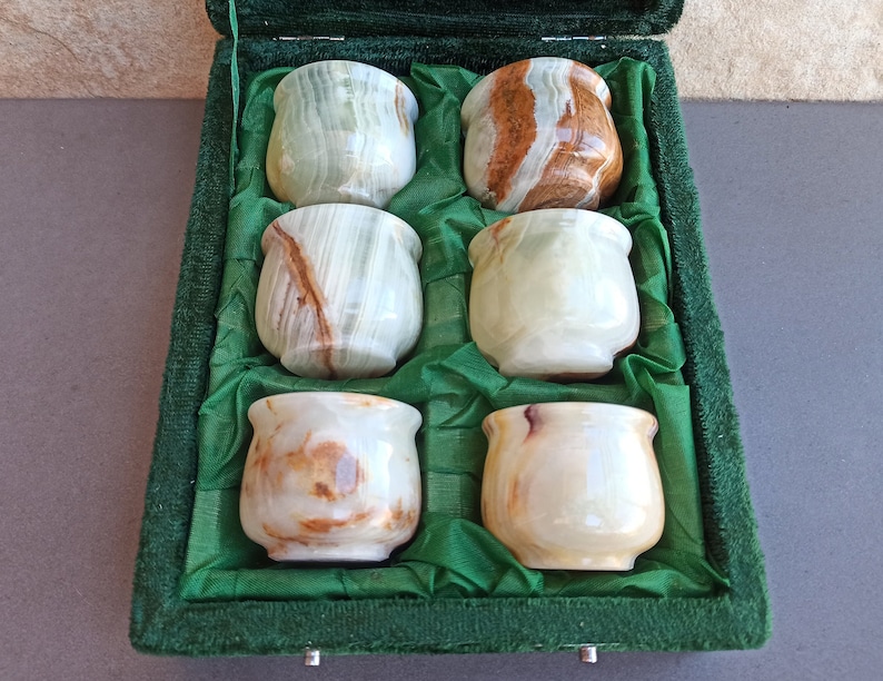 K&ouml;nnte beinhalten: Sechs kleine Onyx-Becher in Gr&uuml;n, Wei&szlig; und Braun sind in einer mit gr&uuml;nem Samt ausgekleideten Schachtel ausgestellt. Die Becher sind in zwei Reihen zu je drei angeordnet.