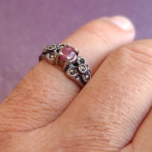 K&ouml;nnte beinhalten: Ein silberner Ring mit einem ovalen, rosafarbenen Edelstein in der Mitte, umgeben von kleinen, dekorativen floralen Akzenten. Der Ring wird an einem Finger getragen und zeigt sein aufwendiges Design und seinen Vintage-Stil. Das Metall des Rings hat ein leicht angelaufenes Aussehen.