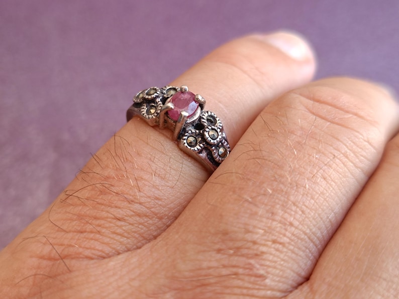 K&ouml;nnte beinhalten: Ein silberner Ring mit einem ovalen, rosafarbenen Edelstein in der Mitte, umgeben von kleinen, dekorativen floralen Akzenten. Der Ring wird an einem Finger getragen und zeigt sein aufwendiges Design und seinen Vintage-Stil. Das Metall des Rings hat ein leicht angelaufenes Aussehen.