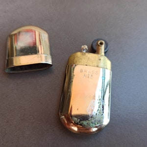 May include: A vintage brass lighter with a black wheel and a lid. The lighter has the words "Brass No. 5" engraved on the side.