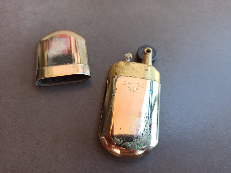 May include: A vintage brass lighter with a black wheel and a lid. The lighter has the words "Brass No. 5" engraved on the side.