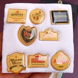 May include: A collection of eight gold-colored metal pins featuring various beer brands. The pins are arranged in two rows on a white surface. The brands include: Murphy's Irish Red Beer, Fischer, Guinness, Vos, Adelscott, Wieckse Witte, Desperados, and Affligem.