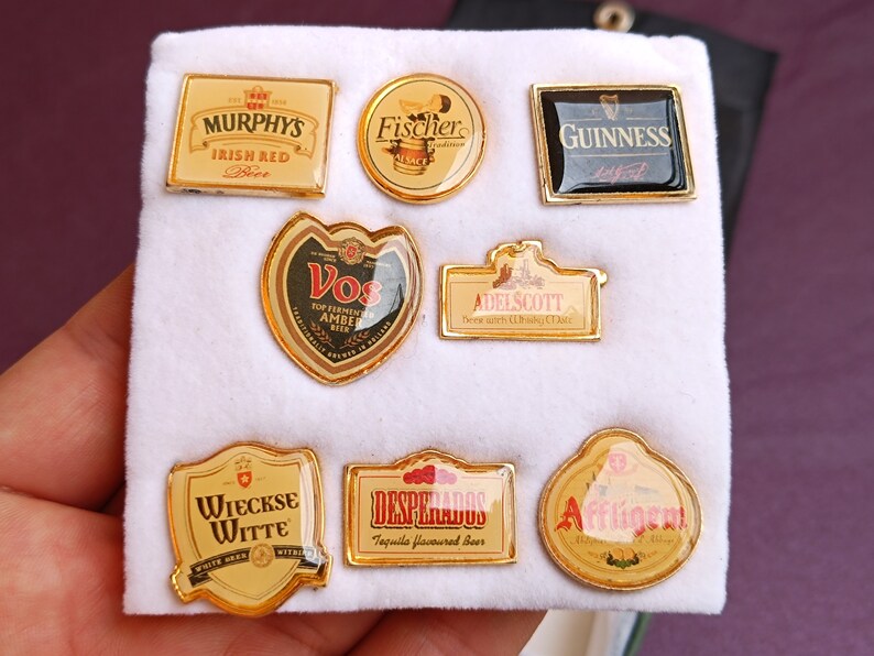 May include: A collection of eight gold-colored metal pins featuring various beer brands. The pins are arranged in two rows on a white surface. The brands include: Murphy's Irish Red Beer, Fischer, Guinness, Vos, Adelscott, Wieckse Witte, Desperados, and Affligem.