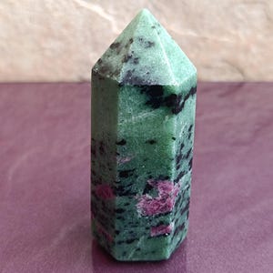 May include: A green and black ruby zoisite crystal point. The polished gemstone has a hexagonal base and a pointed top. Pink ruby inclusions are visible throughout the stone. The crystal is approximately 3 inches tall.