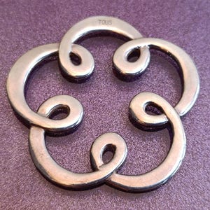 May include: A silver-toned, interlocking jewelry piece with a floral design. The piece is made of curved, polished metal, with the brand name "TOUS" subtly engraved on one of the links. The design is set against a purple background.