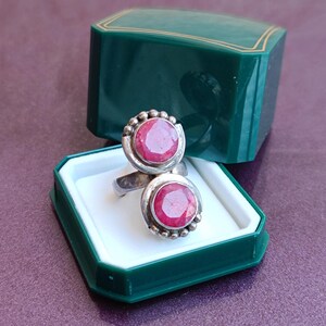 Puede incluir: Un anillo de plata con dos grandes piedras preciosas rojas redondas. El anillo se exhibe en una caja de joyer&iacute;a verde abierta con interior blanco. El fondo es de color p&uacute;rpura s&oacute;lido. El dise&ntilde;o del anillo es llamativo.
