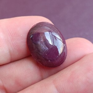 Puede incluir: Una piedra preciosa pulida, de forma ovalada, de color burdeos oscuro. La gema tiene una superficie lisa y reflectante, con sutiles patrones internos. La piedra se sostiene en una mano, con un fondo morado borroso.