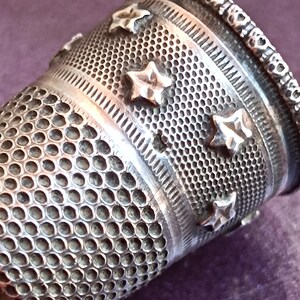 May include: Close-up of a silver thimble with a detailed design. The thimble features a pattern of small holes, star-shaped embellishments, and decorative bands. The top edge has a heart-shaped border.