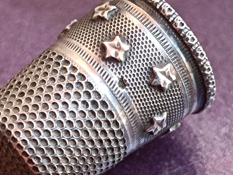 May include: Close-up of a silver thimble with a detailed design. The thimble features a pattern of small holes, star-shaped embellishments, and decorative bands. The top edge has a heart-shaped border.