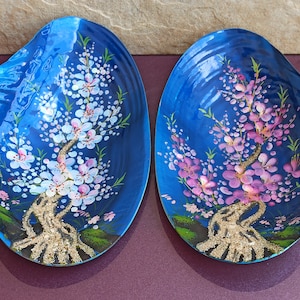 以下が含まれることがあります： ピンクの花と金色のキラキラが描かれた木のデザインが施された青い貝殻が2つ。貝殻は紫色の表面の上に置かれています。