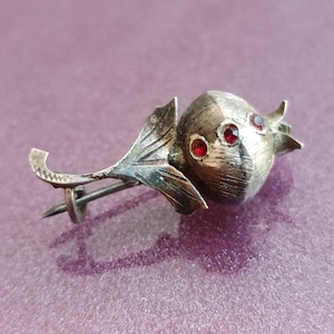 May include: Silver brooch shaped like a pomegranate with three red gemstones. The brooch has an intricate design and a pin on the back.