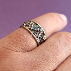May include: A silver-toned metal ring featuring an intricate, raised floral or vine-like pattern across the band. A small, rectangular, dark green gemstone is set centrally within the decorative metalwork on the outer surface of the ring.