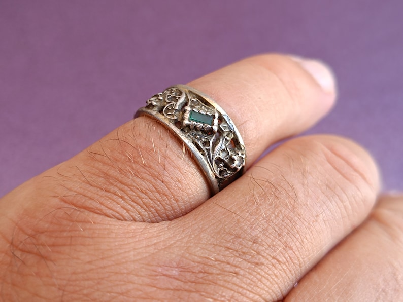 May include: A silver-toned metal ring featuring an intricate, raised floral or vine-like pattern across the band. A small, rectangular, dark green gemstone is set centrally within the decorative metalwork on the outer surface of the ring.