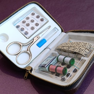 Könnte beinhalten: Eine braune Leder-Nähzeugtasche mit Reißverschluss. Die Tasche ist geöffnet und enthält eine Schere, ein kleines Fläschchen mit Flüssigkeit, zwei Knopfpäckchen, zwei Garnrollen, eine kleine Nadelbox und ein Maßband.