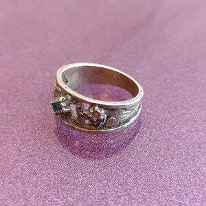 May include: A silver-toned metal ring featuring an openwork band design with intricate filigree detailing. The ring is accented with a small, rectangular green stone set within a decorative metal frame, showcasing a textured and weathered finish.