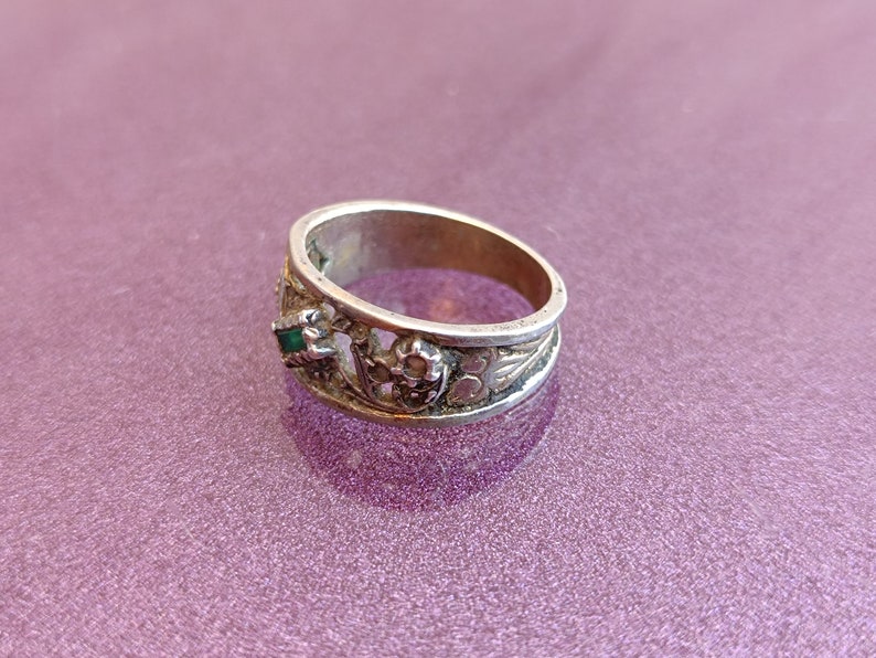 May include: A silver-toned metal ring featuring an openwork band design with intricate filigree detailing. The ring is accented with a small, rectangular green stone set within a decorative metal frame, showcasing a textured and weathered finish.
