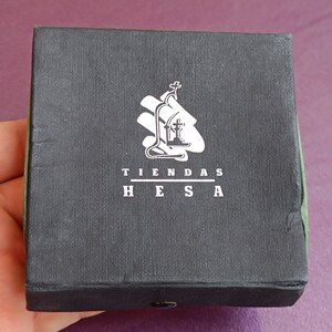 May include: A black box with a white logo that says "TIENDAS HESA". The logo features a stylized image of a church with a cross on top.