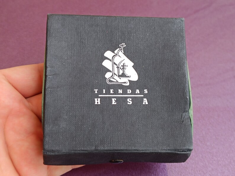 May include: A black box with a white logo that says "TIENDAS HESA". The logo features a stylized image of a church with a cross on top.
