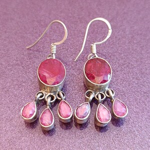 May include: A pair of silver dangle earrings with red gemstone drops. The earrings feature a larger oval-shaped gemstone at the top and three smaller teardrop-shaped gemstones hanging below.