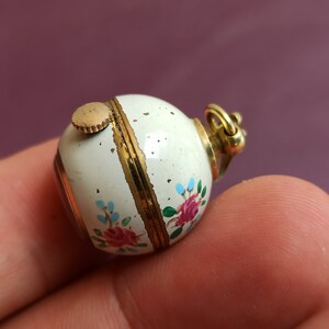 May include: A small, round, white enameled pendant with gold accents. The pendant is decorated with hand-painted pink and red roses and blue floral accents. A gold-colored crown and winding stem are visible.