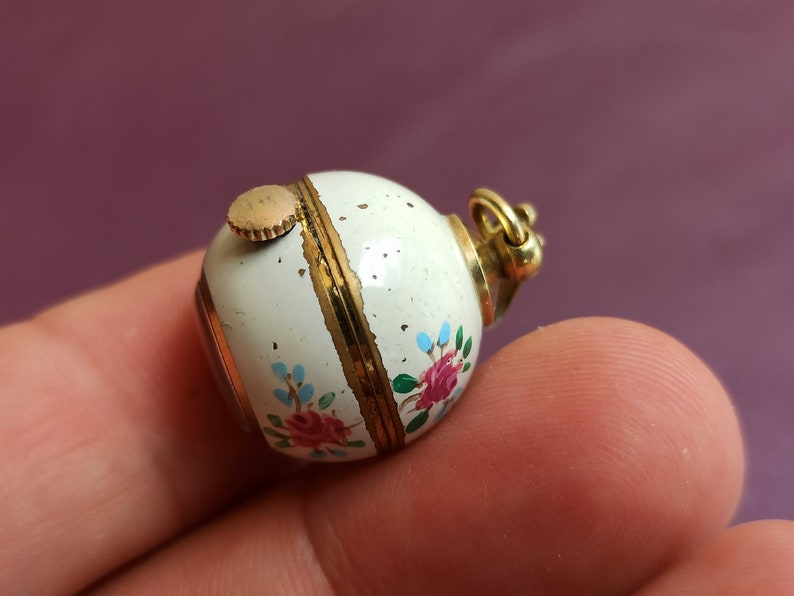 May include: A small, round, white enameled pendant with gold accents. The pendant is decorated with hand-painted pink and red roses and blue floral accents. A gold-colored crown and winding stem are visible.