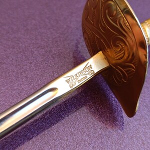 K&ouml;nnte beinhalten: Ein silbernes Schwert mit einem goldfarbenen Schutz und Griff. Das Schwert ist mit den Worten "Wilkinson Sword" graviert.