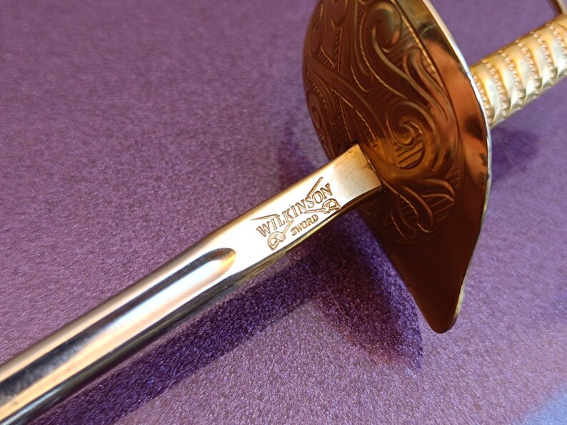 K&ouml;nnte beinhalten: Ein silbernes Schwert mit einem goldfarbenen Schutz und Griff. Das Schwert ist mit den Worten "Wilkinson Sword" graviert.