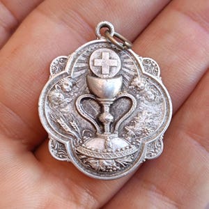 Peut inclure: Un pendentif argenté à thème religieux. Le pendentif représente un calice, une croix et des figures angéliques. Le design est détaillé et orné, avec une esthétique vintage. Le pendentif est probablement en métal et possède une boucle pour l'attacher à une chaîne.