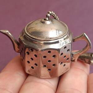 Può includere: Un piccolo infusore per tè color argento a forma di teiera in miniatura. L'infusore ha un coperchio, un beccuccio e un manico, con una catena attaccata al coperchio. Il corpo dell'infusore è perforato con piccoli fori.