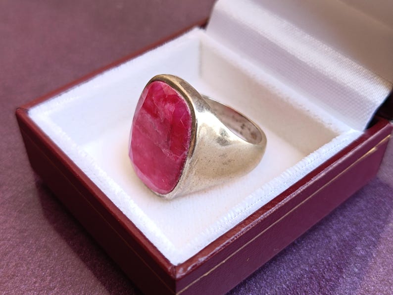 Op de afbeelding: Een zilveren ring met een grote, ovaalvormige, rode edelsteen. De ring wordt gepresenteerd in een rood-witte sieradendoos. De edelsteen heeft een textuur en is het middelpunt van de ring.