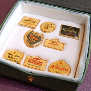 May include: A collection of eight different beer brand pin badges in a green velvet box. The badges are gold-colored and feature the logos of various beer brands, including Murphys Irish Red, Fischer, Guinness, Vos Amber, Adelscott, Wieckse Witte, Desperados Tequila Flavored Beer, and Affligem.