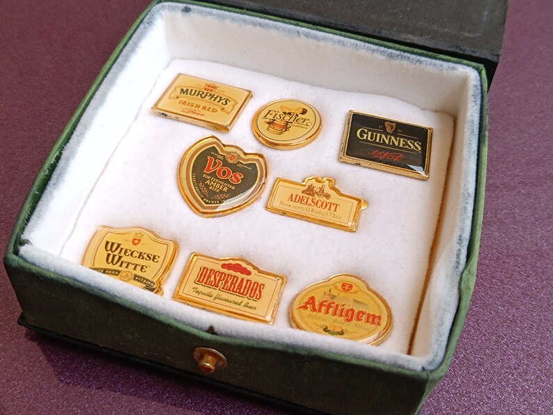 May include: A collection of eight different beer brand pin badges in a green velvet box. The badges are gold-colored and feature the logos of various beer brands, including Murphys Irish Red, Fischer, Guinness, Vos Amber, Adelscott, Wieckse Witte, Desperados Tequila Flavored Beer, and Affligem.
