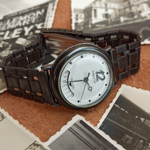 Peut inclure: Une montre-bracelet noire avec un cadran argenté et des aiguilles noires. Le cadran de la montre porte l'inscription "PAKETA" et le chiffre "12". La montre est sur un bracelet en métal noir.
